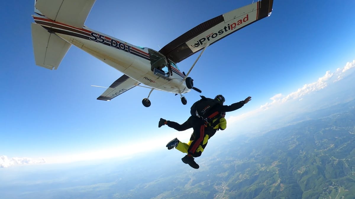 Tandem skydive pair exiting a white aircraft over scenic Slovenian landscape during a clear day.