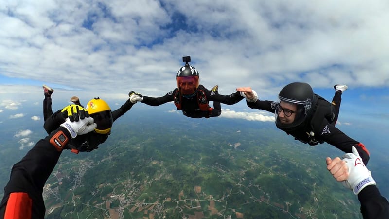 Four skydivers in formation during freefall, holding hands in a circle high above a landscape of fields and hills under a partly cloudy sky. They wear colorful helmets and jumpsuits, with one skydiver using a helmet-mounted camera.