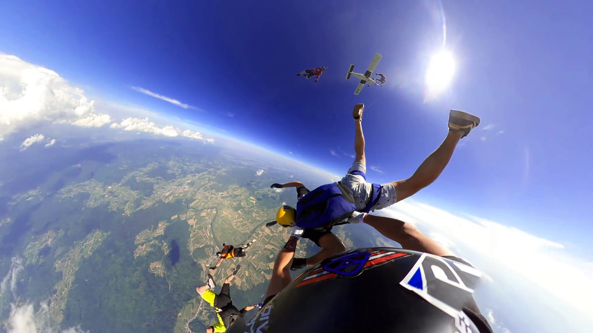 Group skydive in freefall with blue skies, plane overhead, and scenic Slovenian landscape below