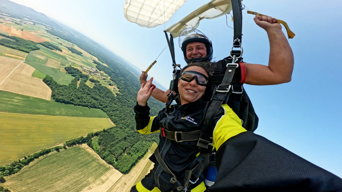 Tandem skydiving pair smiling mid-air with open parachute above scenic fields and forests in clear blue sky