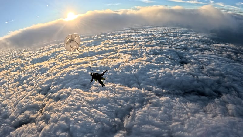 Skydiver descending under parachute during a sunset skydive above the clouds in Slovenia, with golden light illuminating the sky and cloud layer below.