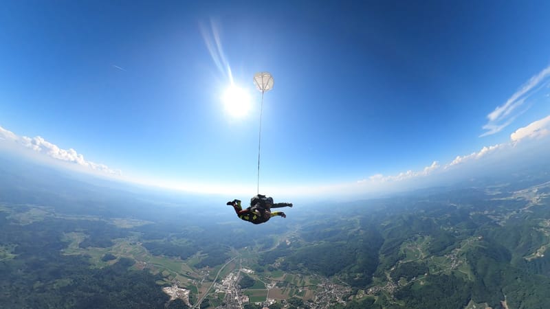 Skydivers in free fall with drogue chute deployed above scenic landscape in Slovenia.